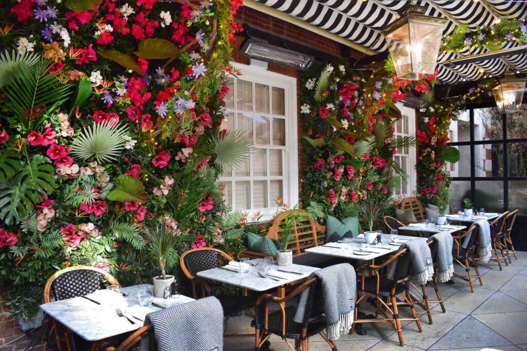 Adorable outdoor restaurant with bright flowered walls and blankets over wicker chairs to keep patrons warm along with a Bromic Heater